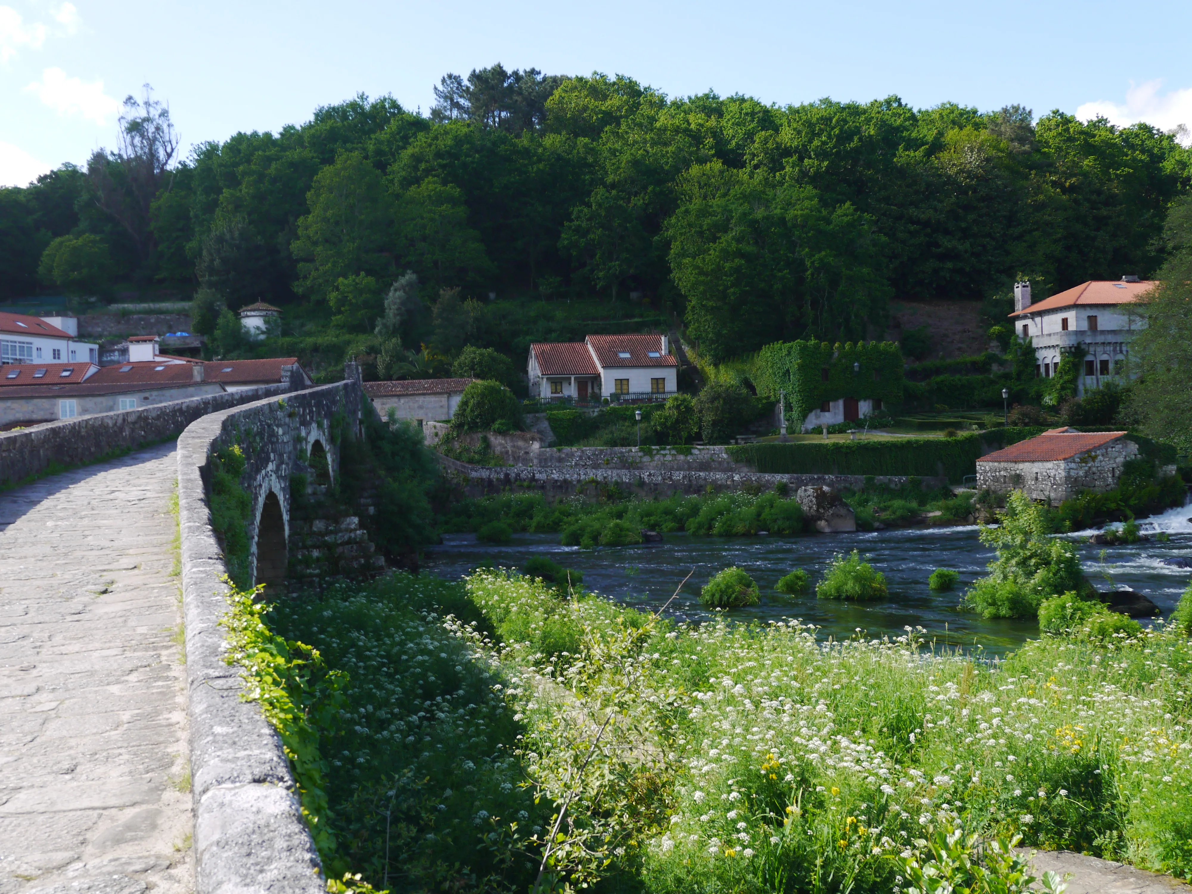 A historic stone bridge crossing a river beside houses and woodland in Galicia.