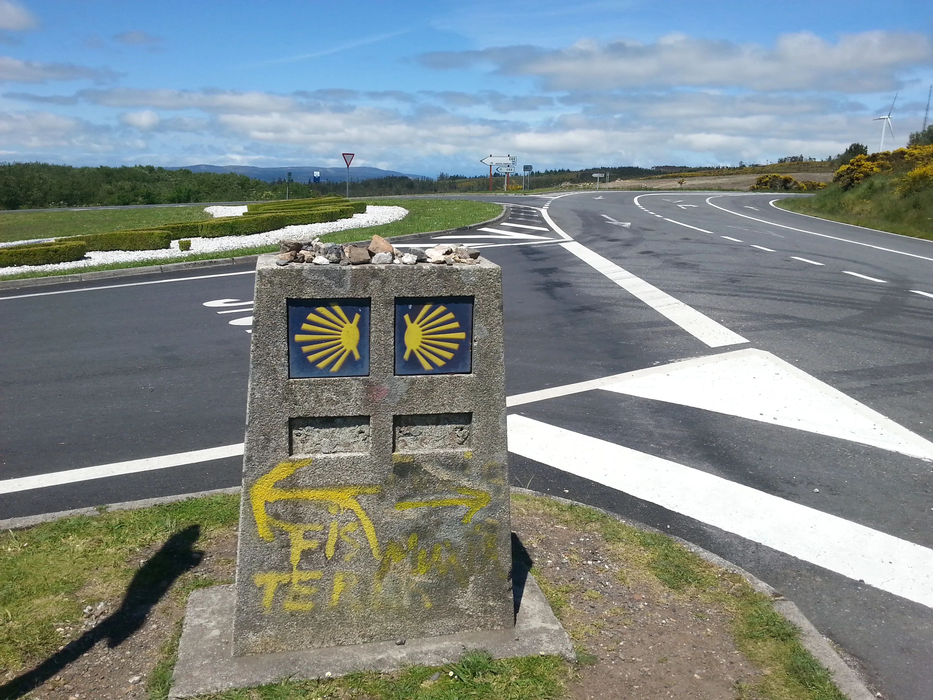 A Camino waymarker at a junction, with yellow arrows pointing in different directions.